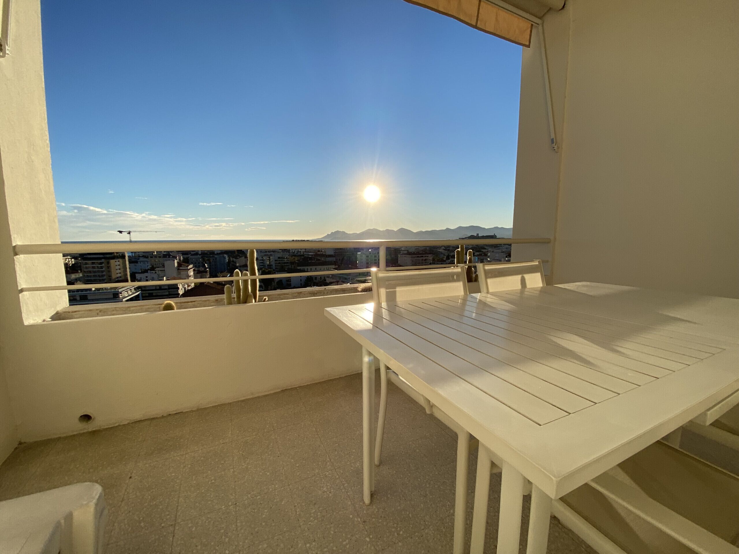 A balcony with a white table and chairs, a view of the sea and mountains, and a clear sky.