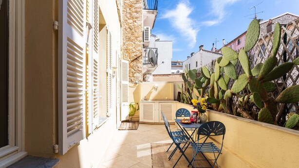 Un patio soleado con una mesa y sillas, un muro de cactus y un edificio con persianas.