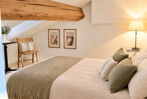 A bedroom with a bed, pillows, a lamp, a chair, and two framed pictures on the wall.