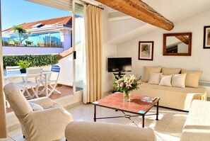 A modern living room with a sofa, armchairs, a coffee table, and a TV. There is a balcony with a table and chairs, and a view of a building with a tiled roof.