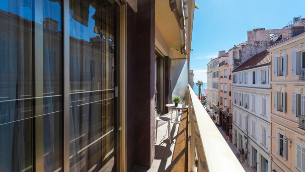 A balcony with a view of a narrow street lined with buildings, a small table with a plant, and a railing. - Appartement Trois pièces DEB5515 (Cannes)