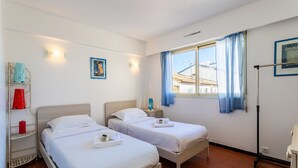 A room with two beds, a window with blue curtains, a shelf with red and blue items, and a wall-mounted picture. - Appartement Trois pièces DEB5515 (Cannes)