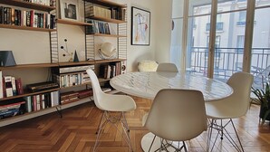 A modern living room with a round dining table, white chairs, a bookshelf, and a large window with a view of buildings. - Riviera Design / Charming apartment 2mn from the beach (Cannes)