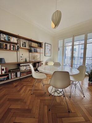 A modern living room with a round dining table, white chairs, a bookshelf, and a large window with a view of buildings. - Riviera Design / Charming apartment 2mn from the beach (Cannes)