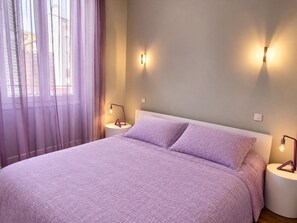 A bedroom with a bed, two bedside lamps, a window with sheer curtains, and a wall-mounted light fixture.