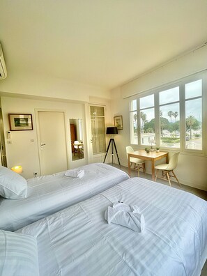 Een hotelkamer met twee bedden, een bureau, een stoel en een raam met uitzicht op palmbomen.