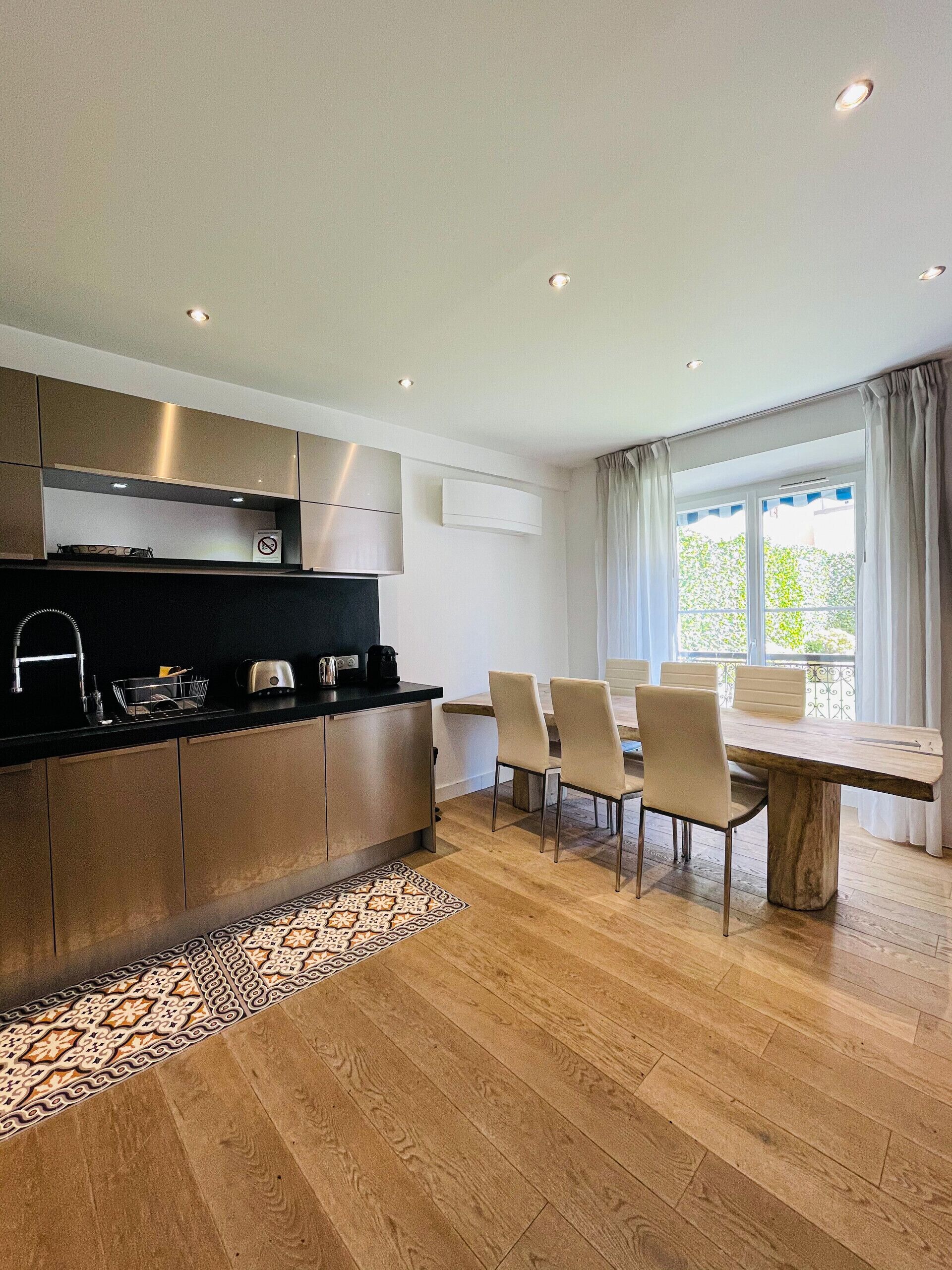 A modern kitchen with a dining area, wooden flooring, and a view through a window.