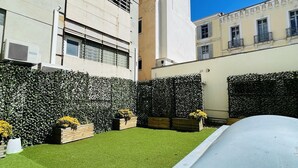 Un patio con césped artificial, un coche y un edificio de fachada blanca y numerosas ventanas.