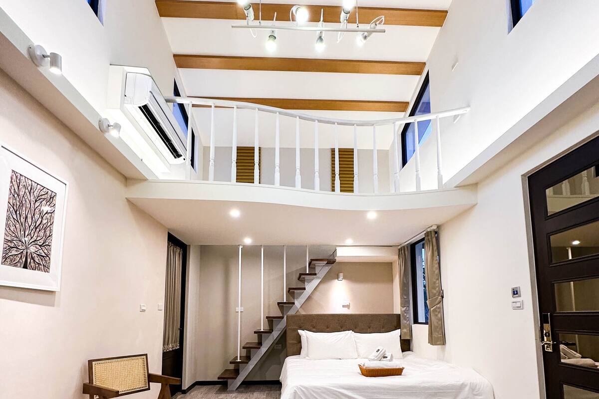 A modern hotel room with a loft-style design, featuring a bed, a seating area, and a staircase leading to an upper level.