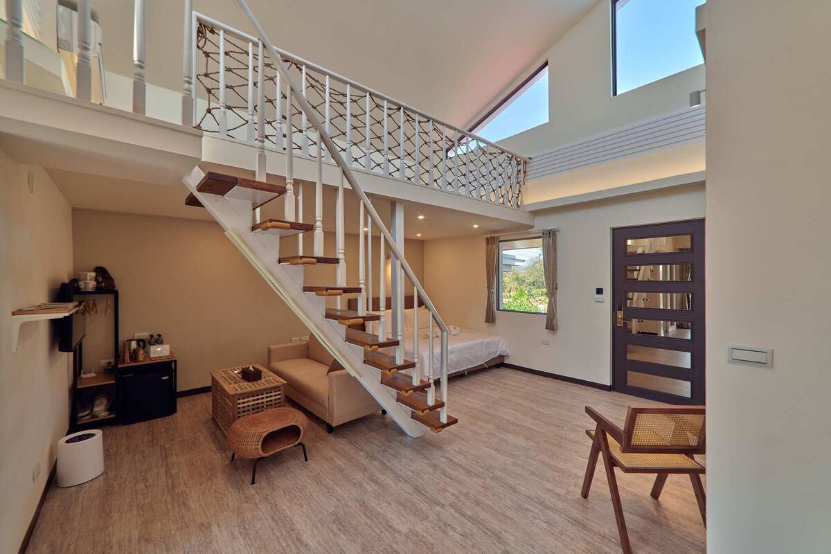 A spacious living area with a modern staircase, a sofa, a wicker chair, and a bookshelf.