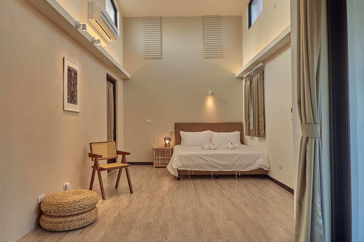 A bedroom with a bed, a chair, a bedside table, and a wall-mounted air conditioner.