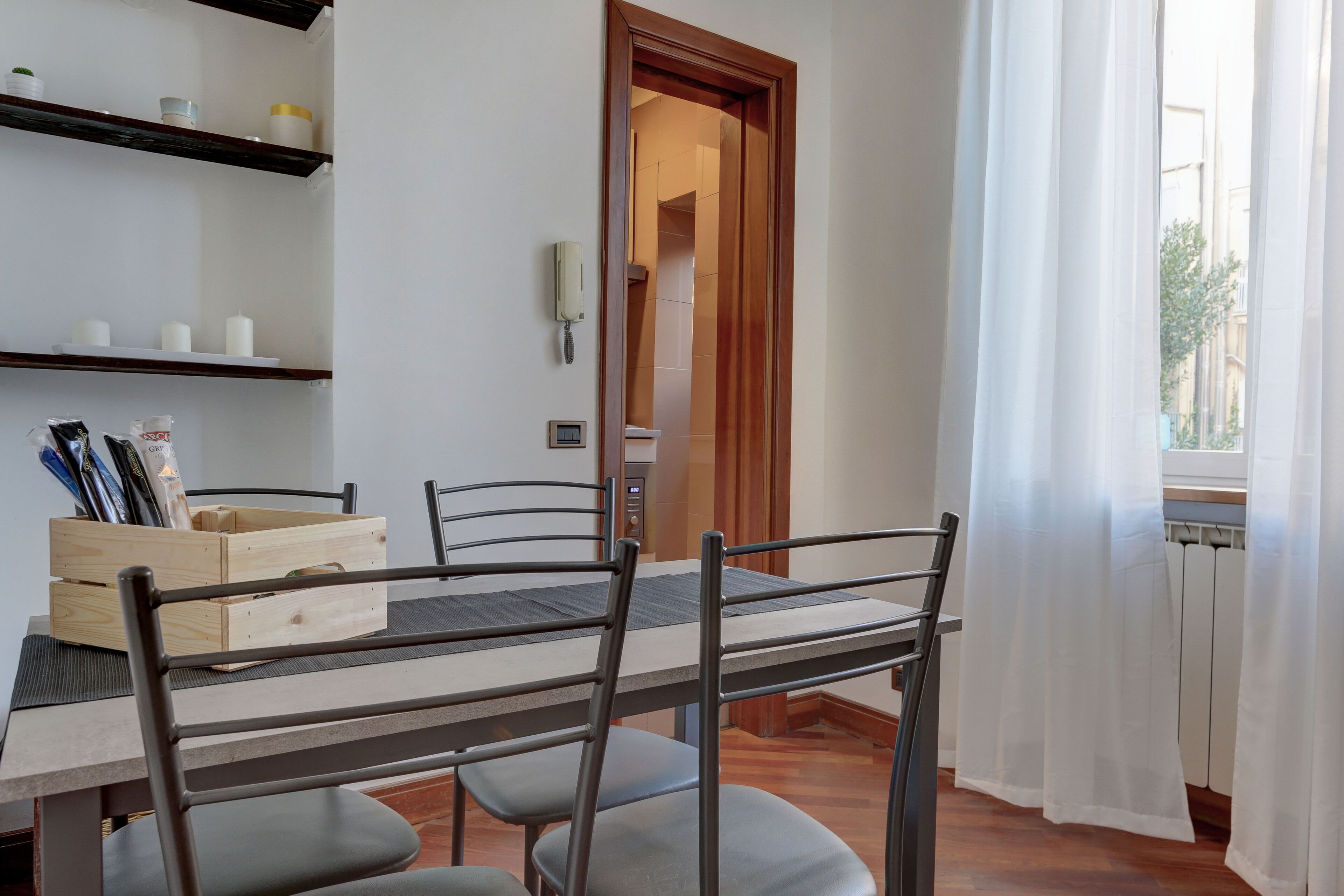 A dining area with a wooden table, metal chairs, and a wooden crate. There is a white wall with shelves holding various items. A door leads to another room.