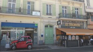 Straßenansicht mit einem roten Auto vor einem Gebäude, dessen grüne Tür und Schild mit dem Schriftzug "LE TRIOMPHE" zu sehen sind.