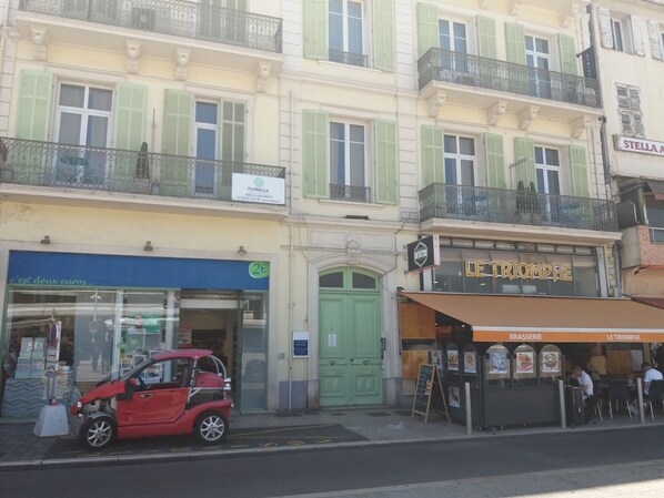 A street view with a red car parked in front of a building with a green door and a sign that reads "LE TRIOMPHE".