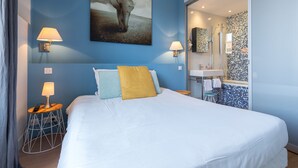 Ein modernes Hotelzimmer mit einem großen Bett, zwei Nachttischlampen, einem Elefantenbild, einem Badezimmer mit gefliesten Wänden und einer Glastür zum Duschbereich.