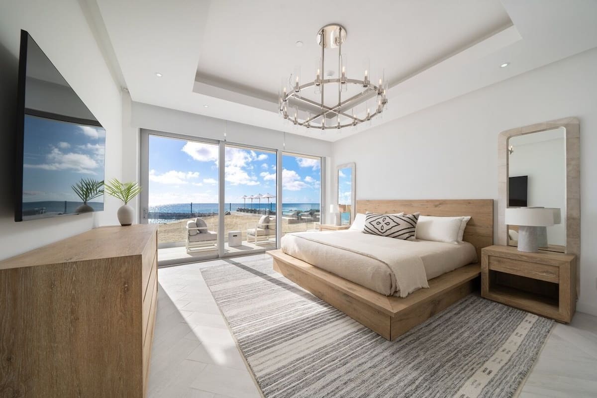 Een moderne slaapkamer met een groot bed, een houten nachtkastje, een grote spiegel en uitzicht op het strand door ramen van vloer tot plafond.