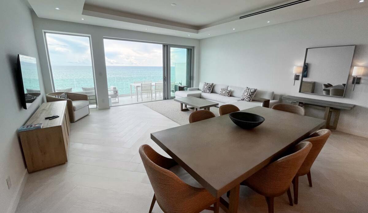 Una habitación de hotel moderna con una gran mesa de comedor, asientos cómodos, televisión y un balcón con vista al mar.