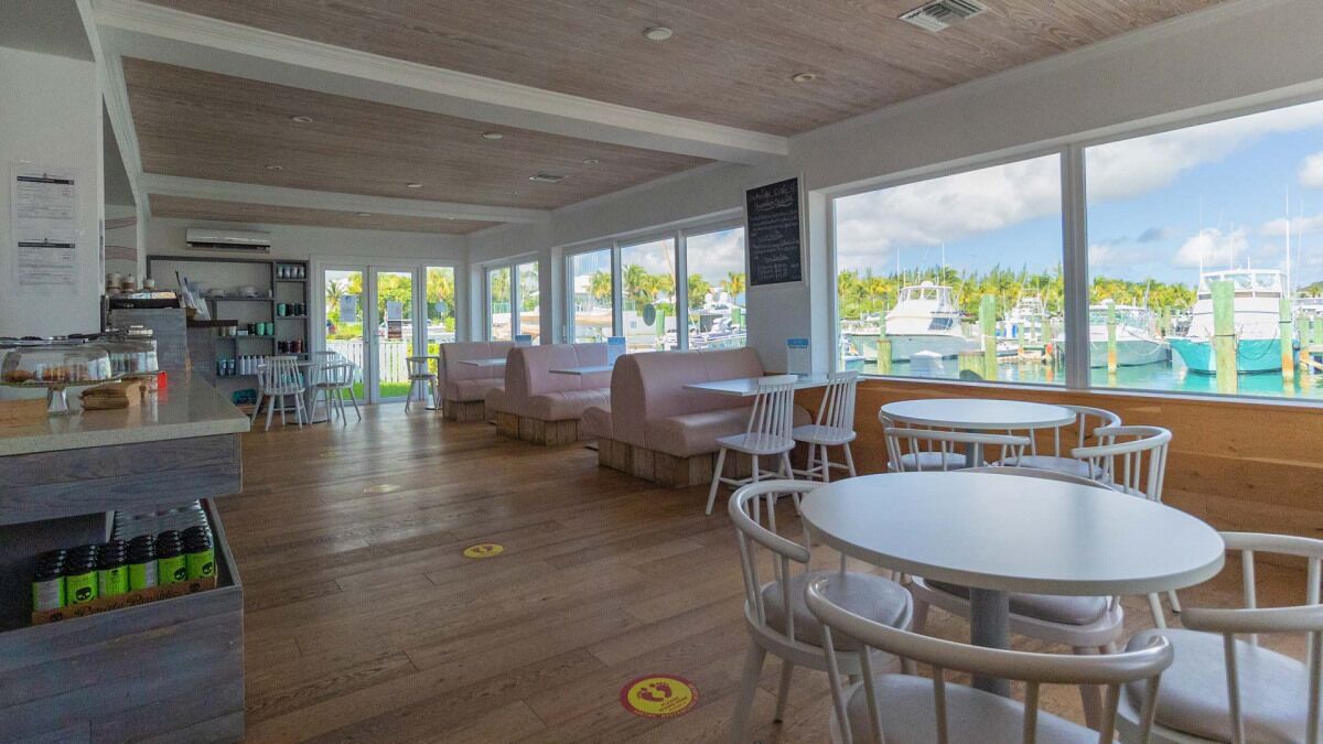 A modern restaurant with wooden floors, white furniture, and large windows offering a marina view.