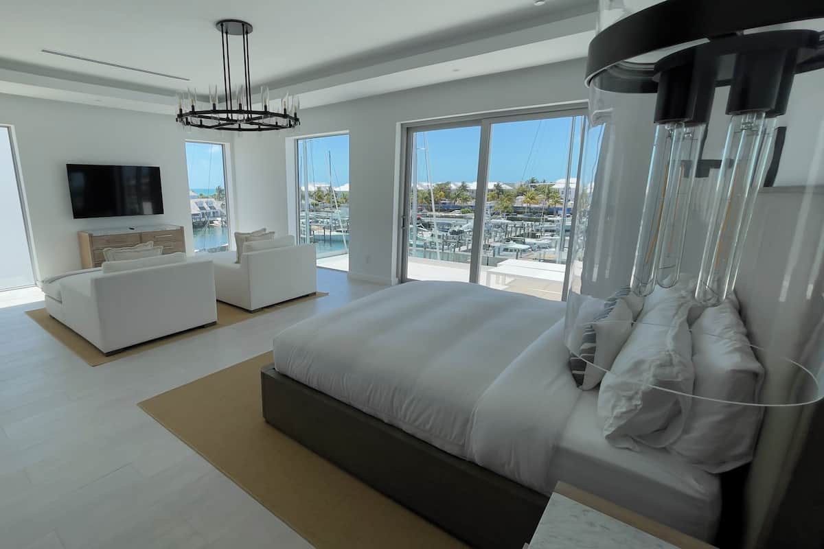 Een moderne slaapkamer met een groot bed, een bank, een televisie en een balkon met uitzicht op boten en palmbomen.