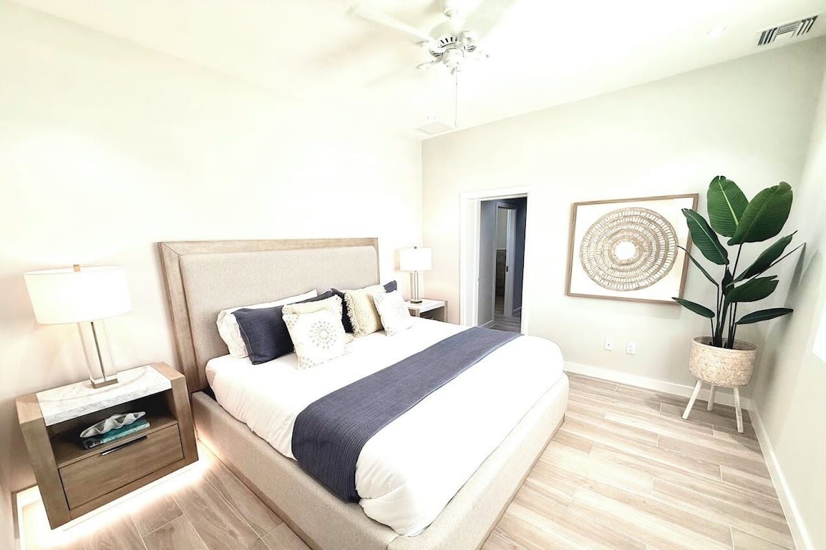 Een moderne slaapkamer met een groot bed, nachtkastjes, een plafondventilator, een potplant en een lijst met kunst aan de muur.