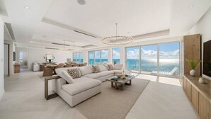Un salon spacieux avec un grand canapé d’angle, une table basse et un téléviseur posé sur un meuble en bois. La pièce est dotée de grandes fenêtres qui offrent une vue sur l’océan.