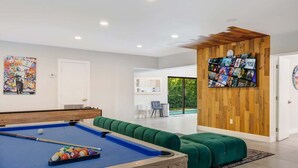 A modern living room with a pool table, green seating, a wooden wall with a TV, and a door leading to a balcony.