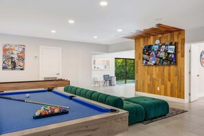 A modern living room with a pool table, green seating, a wooden wall with a TV, and a door leading to a balcony. - Swish Villa | Unique BKB Court | Outdoor Cinema (Miami)
