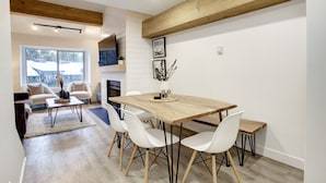 Een moderne eetruimte met een houten tafel en witte stoelen, een open haard en een groot raam met uitzicht op een besneeerd landschap.