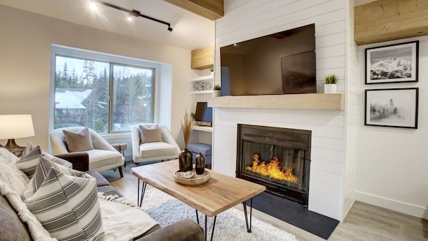 Een moderne woonkamer met een open haard, comfortabele zitplaatsen en een groot raam met uitzicht op bomen.