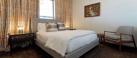 Un dormitorio con una cama, una silla, un cuadro en la pared y cortinas.