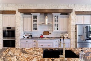 Una cocina con armarios blancos, una pared de piedra, horno empotrado, refrigerador de acero inoxidable y encimera de granito.
