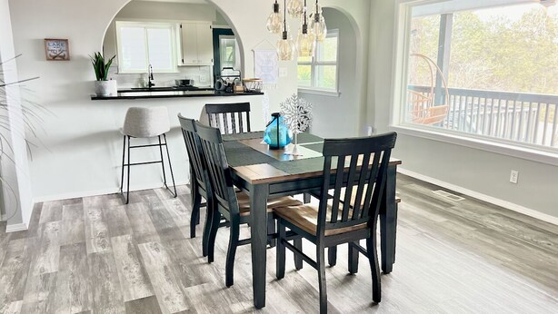 Een eetruimte met een houten tafel en stoelen, een keuken op de achtergrond, en een groot raam met uitzicht.