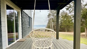 Een witte macramé hangstoel op een houten terras met een overdekte veranda, met uitzicht op een gazon met bomen.