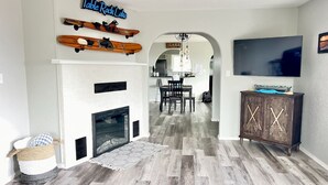 Een woonkamer met een open haard, houten vloer, een televisie en een eethoek op de achtergrond.