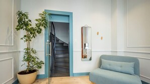 Un pasillo con una planta en maceta, una puerta azulada que da a una escalera, un espejo en la pared y un sofá azul claro.
