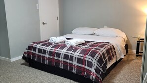Una cama bien hecha con colcha escocesa y dos almohadas, una mesita de noche con lámpara y una puerta blanca.