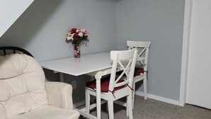 Una habitación pequeña con una mesa blanca y dos sillas blancas con cojines rojos. Un jarrón con flores está sobre la mesa.