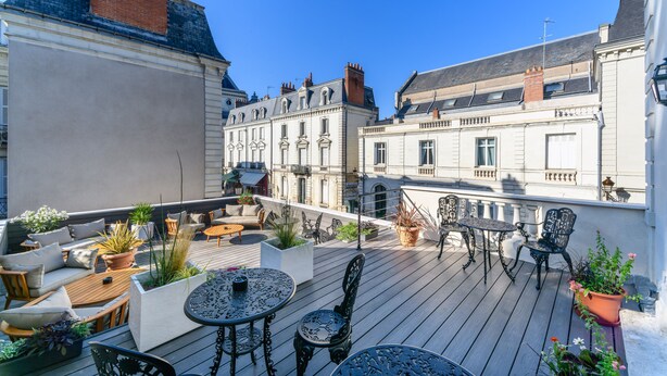 Een terras op het dak met buitenmeubilair, plantenbakken en uitzicht op historische gebouwen.