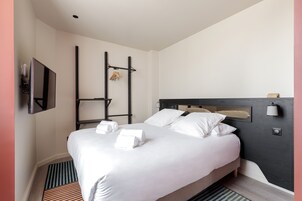 Una habitación de hotel moderna con una cama, un televisor de pantalla plana, un cabecero de madera y un perchero de toallas en la pared.