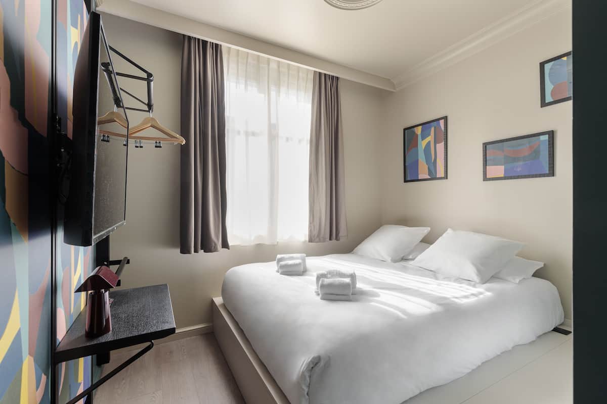 Ein Hotelzimmer mit einem Bett, einem an der Wand befestigten Fernseher, einem Schreibtisch mit Stuhl und einem fensterbelaubten Fenster.