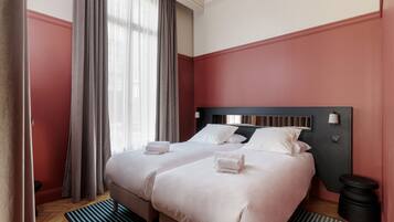 A hotel room with two beds, a wooden floor, a striped rug, a black trash bin, and a window with curtains.