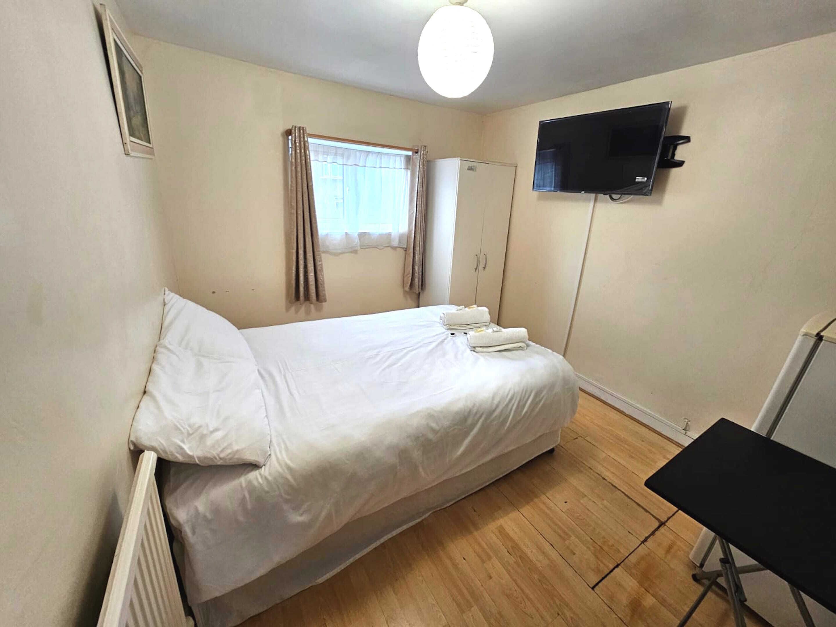 A single bed with white linens, a small window with curtains, a flat-screen TV mounted on the wall, and a radiator on the floor.