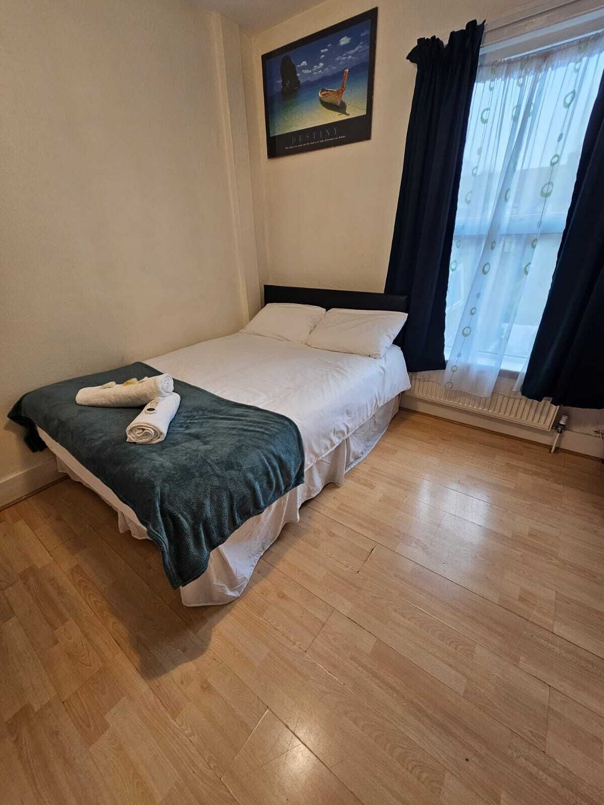 En pænt redt seng med et grønt tæppe og sammenrullede håndklæder, et vindue med lette gardiner, og et indrammet billede på væggen.