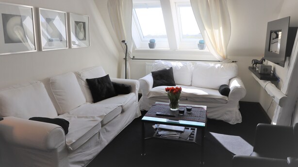 Un salon moderne avec deux canapés blancs, une table basse en verre et une petite table d’appoint. On peut y voir des tableaux encadrés au mur, une plante en pot et une fenêtre avec des rideaux.