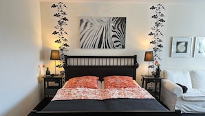 Une chambre avec une tête de lit noire, deux tables de chevet avec des lampes, un lit avec une literie à motifs rouges et blancs, et un canapé blanc.