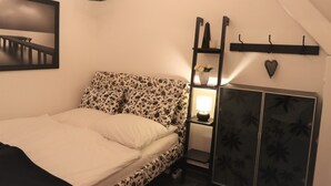 Une chambre à coucher avec un lit, une table de chevet avec une lampe, une étagère avec des fleurs, un porte-manteau et un tableau encadré au mur.