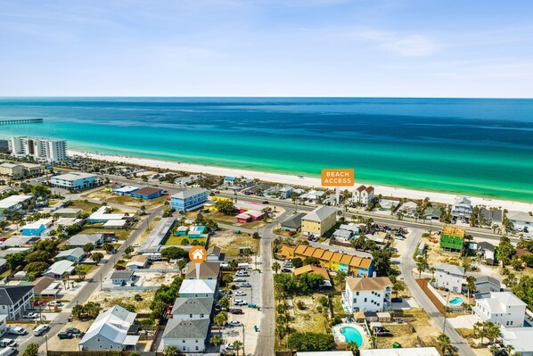 An aerial view of a coastal town with a beach access point, featuring a mix of residential and commercial buildings, roads, and a pier in the distance.