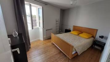 Een slaapkamer met een houten vloer, een bed met houten hoofdbord, nachtkastje, een lamp en een raam met gordijnen.