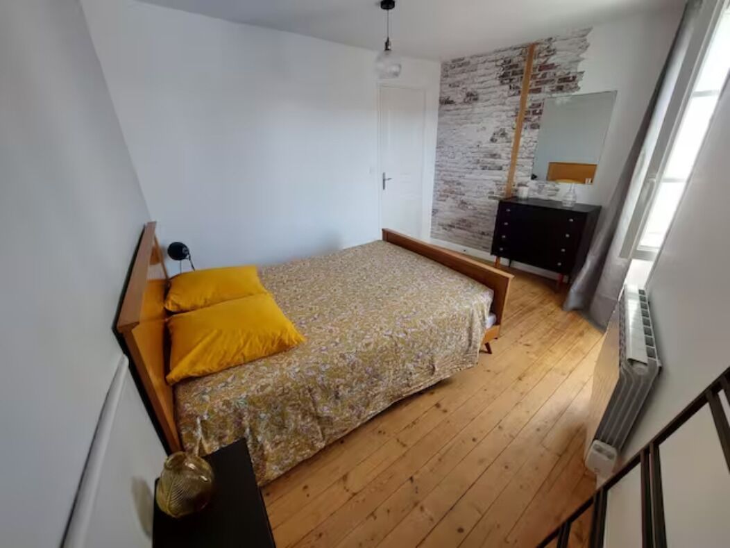 Una camera da letto con pavimento in legno, un letto con copriletto floreale e cuscini gialli, un comò e una parete con motivo a mattoni.
