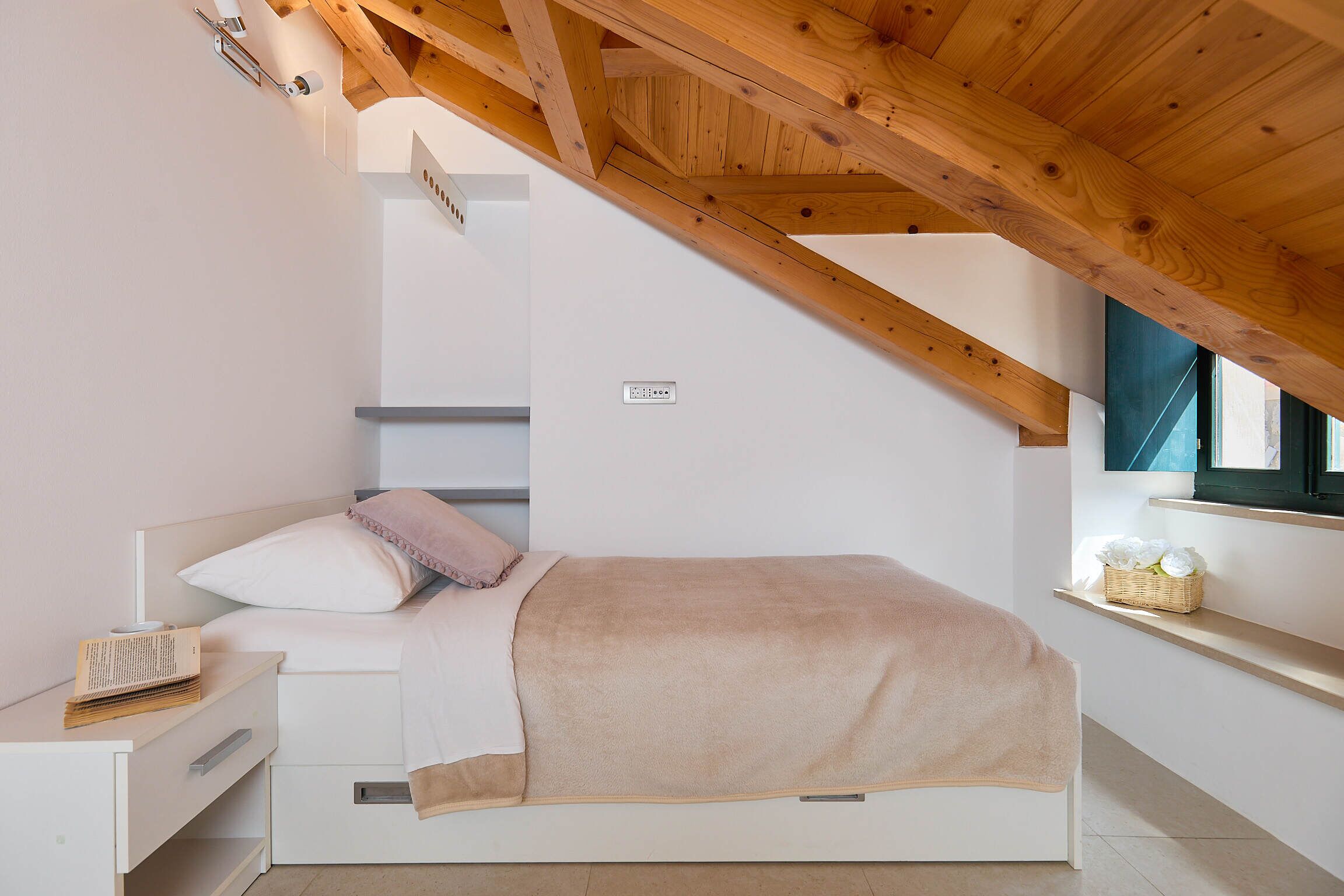 A bedroom with a sloped ceiling, a bed with a beige blanket, a bedside table with a book, and a window with a view.